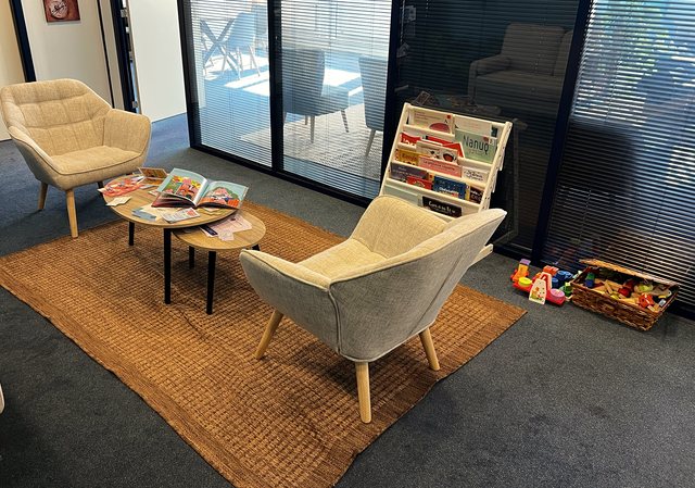 La salle d'attente : deux fauteuils clairs sur un tapis, une table basse avec un livre d'enfant ouvert, une bibliothèque de livres pour enfants et un panier de jeux en bois