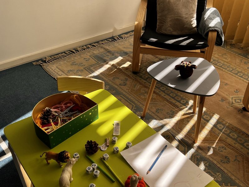 La pièce de consultation au soleil : au premier plan, une petite table verte avec une boîte à trésors, des pommes de pin, des figurines, des dés à histoires, des crayons et un dessin ; derrière, un fauteuil et une petite table avec une chouette en laine